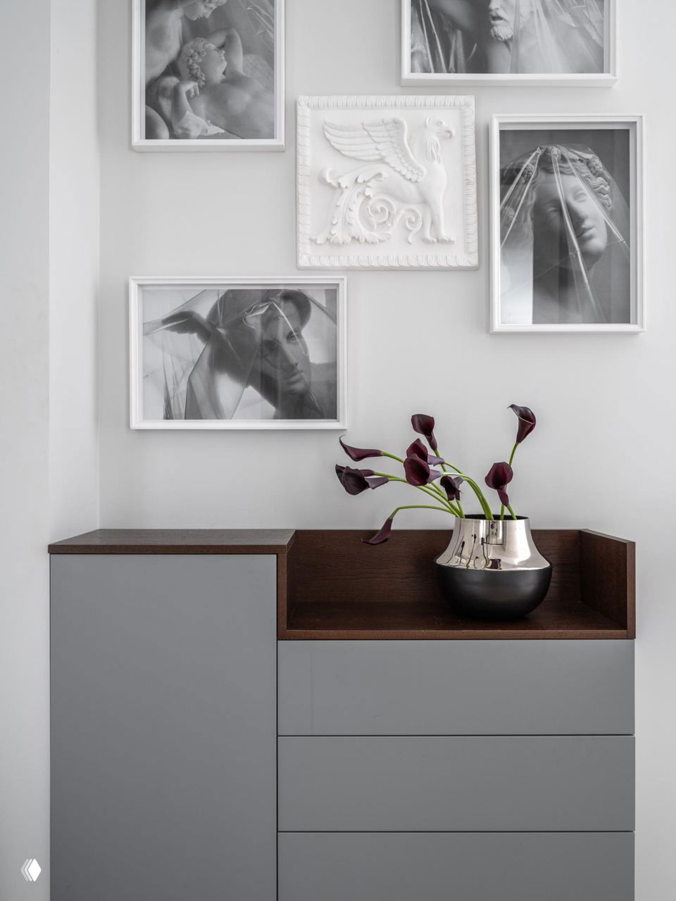 Декор и комод: светло-серая стена с галереей чёрно‑белых фотографий и декоративным барельефом, вазочка с цветами на столе.