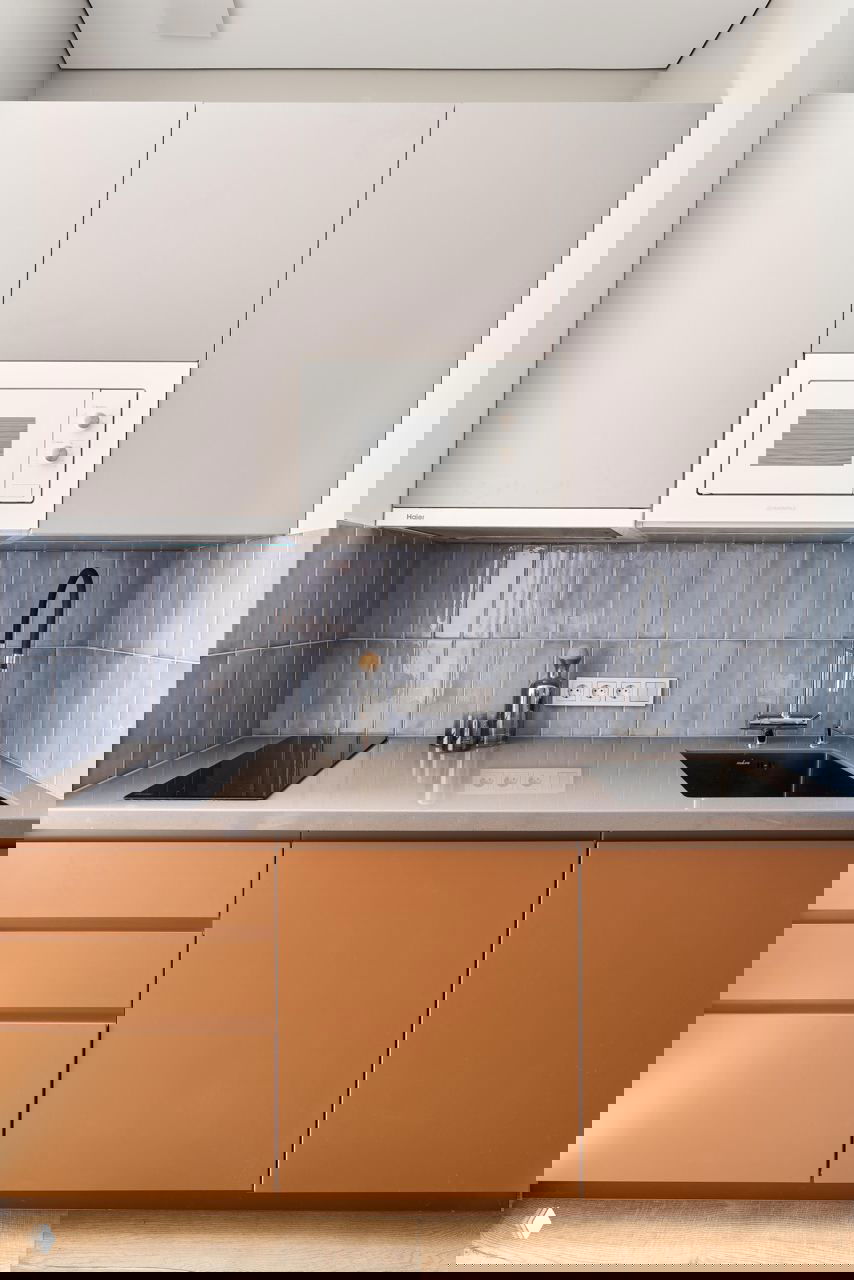 Кухонный гарнитур с матовыми фасадами, голубой плиточный фартук, встроенная микроволновка и мойка с высоким черным смесителем.