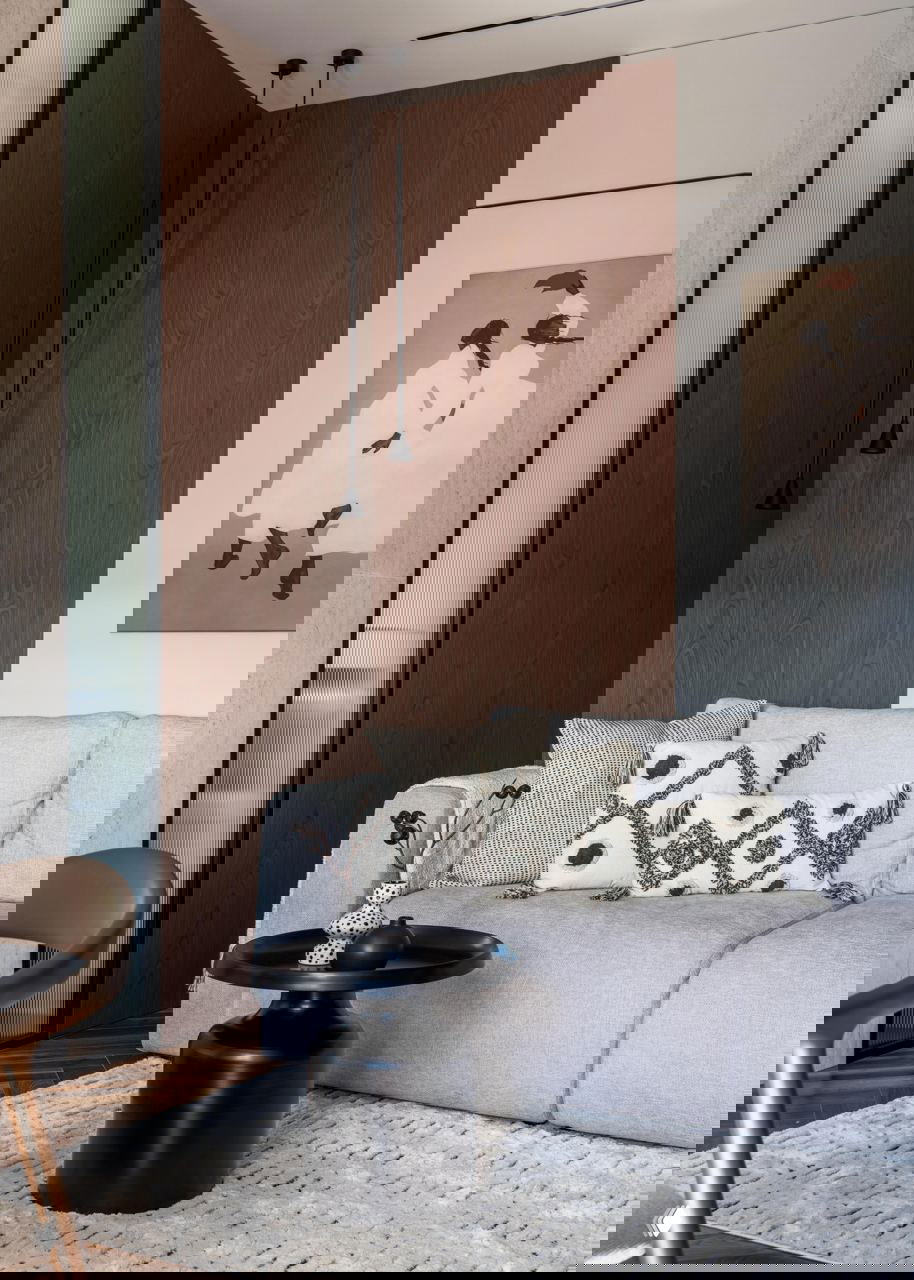 Крупный план дивана с текстурированными подушками, деревянная стеновая панель и два подвесных чёрных светильника в углу, композиция в нейтральной палитре.