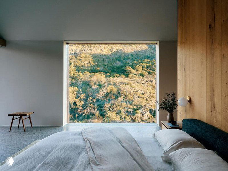 Спальня с минималистичным решением: большая кровать, светлые текстуры и широкое окно во всю стену, через которое видны холмы и природа.