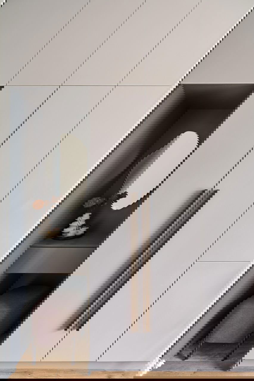 Встроенный светло‑серый гарнитур с маленькой нишей и зеркалом, рядом бархатный пуф — продуманный уголок в компактной квартире.