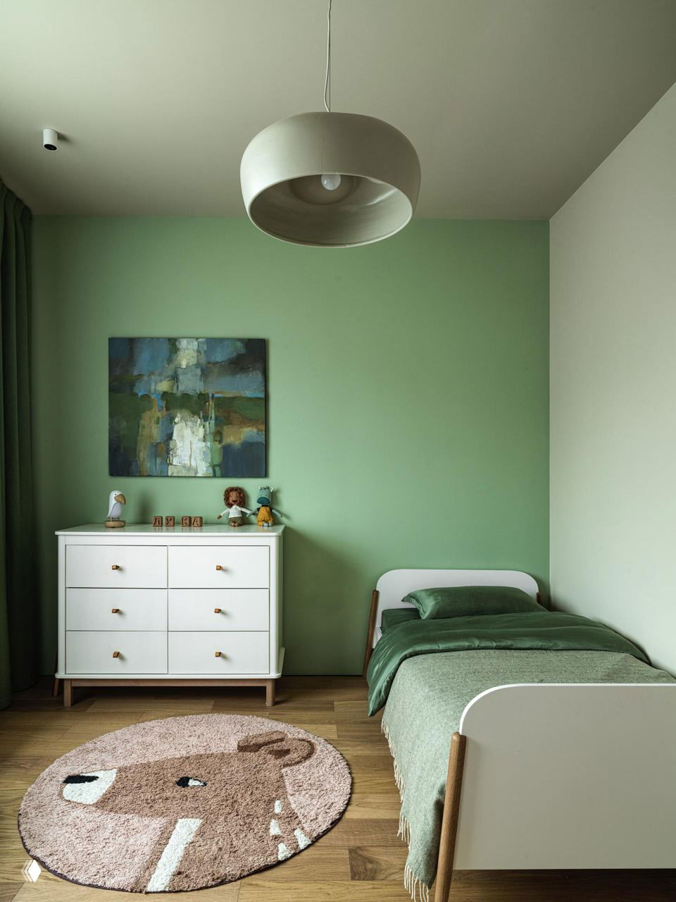Детская комната с нежно-зелёной стеной, односпальной кроватью, белым комодом и круглым ковриком с рисунком животного; естественные деревянные полы.