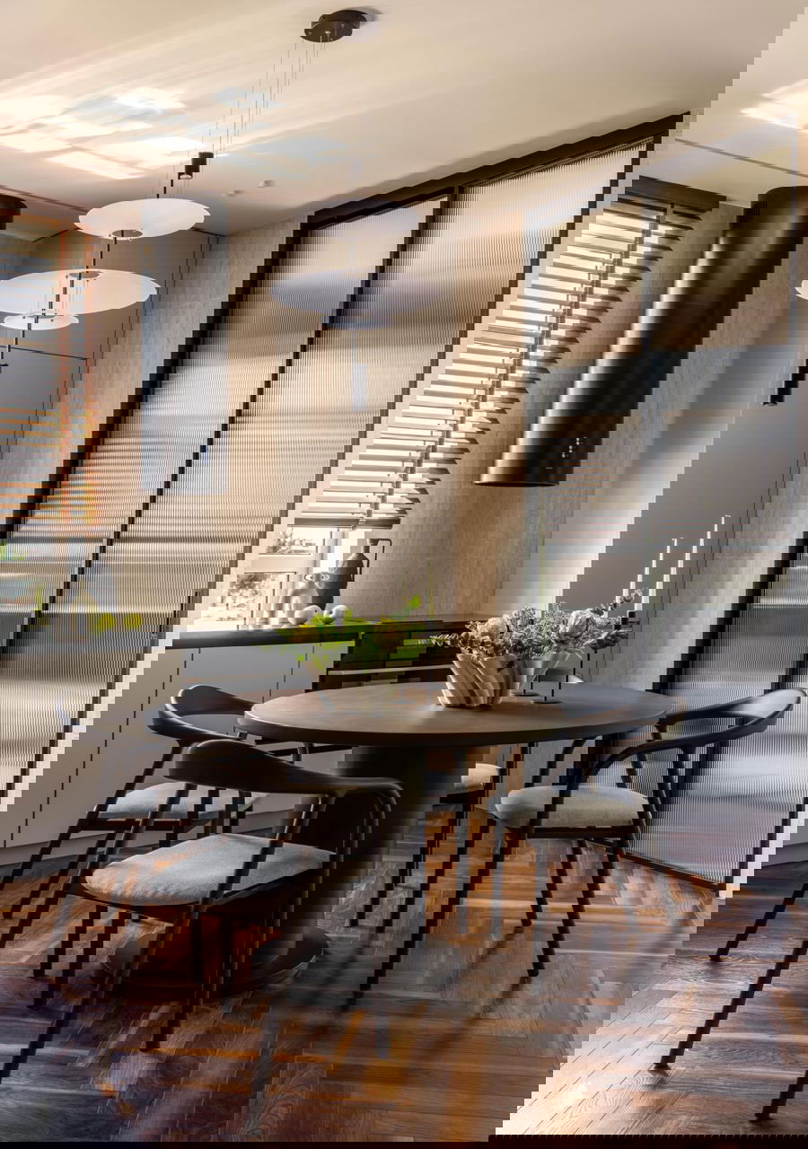 Кухня-столовая с круглый столом, тёмными стульями, вытяжкой цилиндрической формы и рифлёными стеклянными дверями, солнечный свет на паркетном полу.