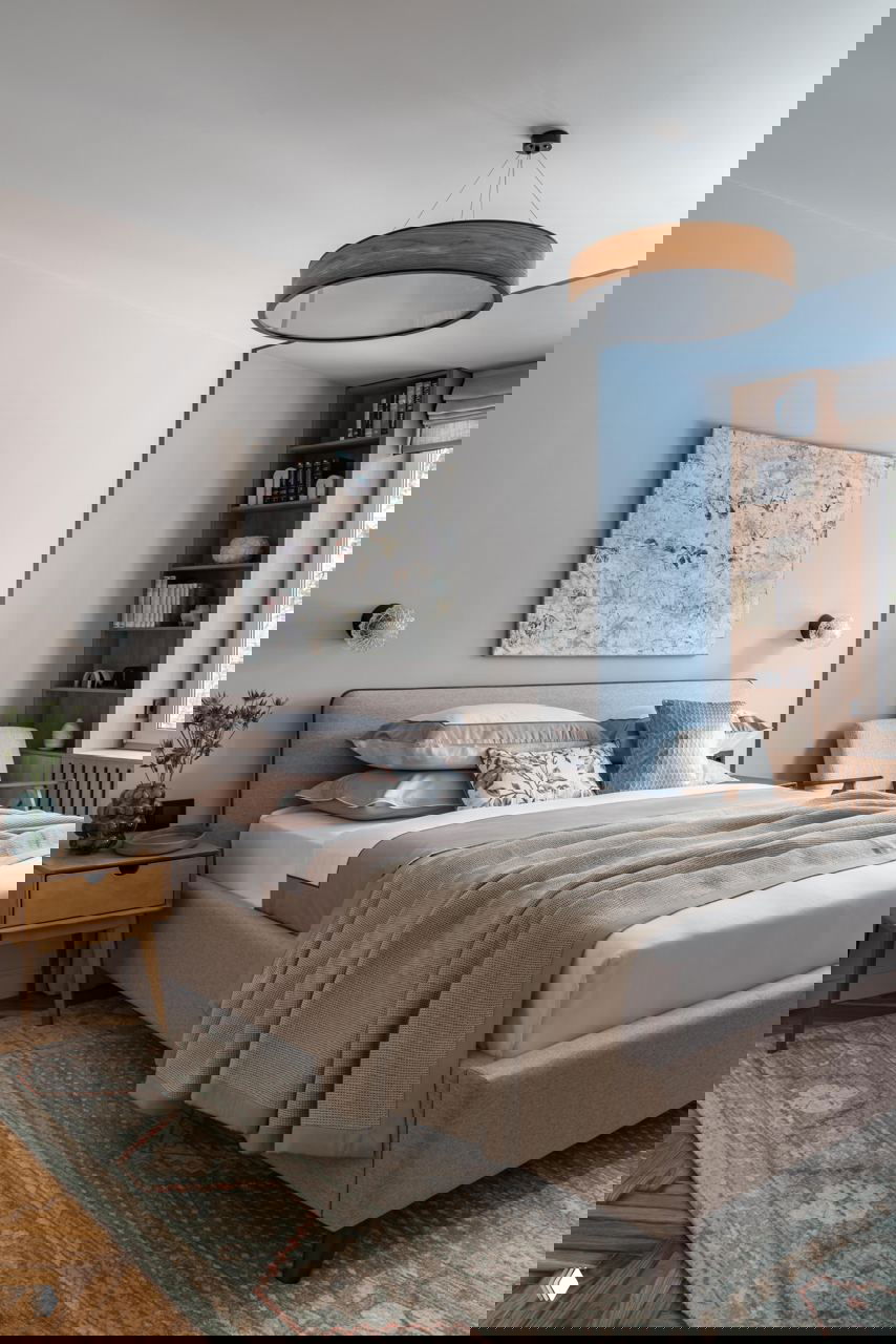 Спальня в спокойной пастельной палитре: мягкая кровать, прикроватная тумба, текстиль и книжный стеллаж в нише — уют и функциональность в одной комнате.