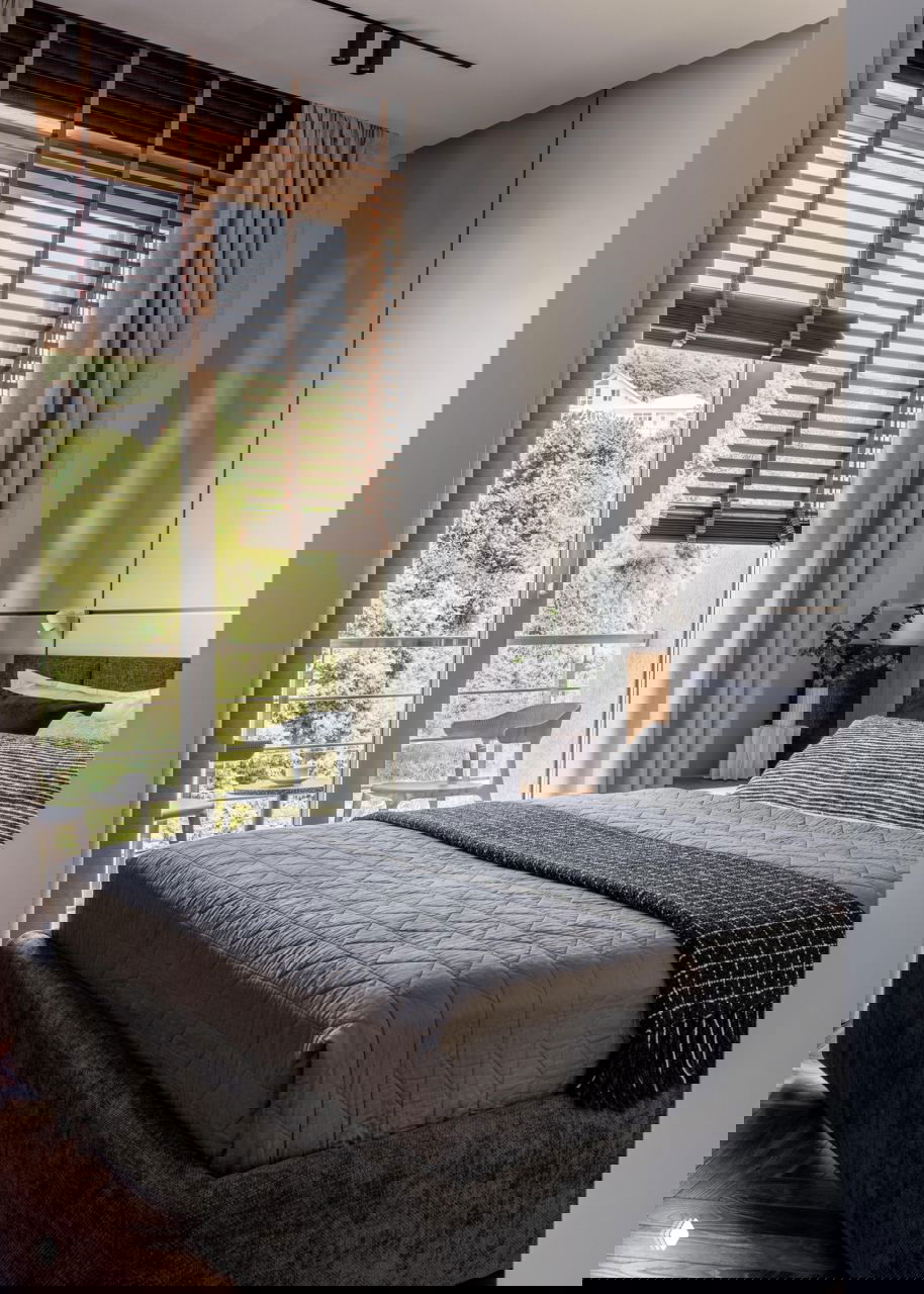 Спальня с большим окном и деревянными жалюзи, кровать в сдержанной серой гамме, текстиль и вид зелёного участка за окном, продуманное естественное освещение.