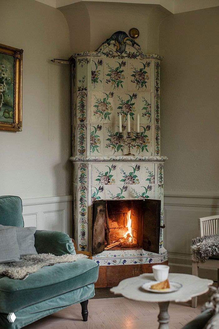 Уютный интерьер со светлой расписной изразцовой печью, горящим огнём в топке, мягким креслом и маленьким кофейным столиком с чашкой и пирожным.