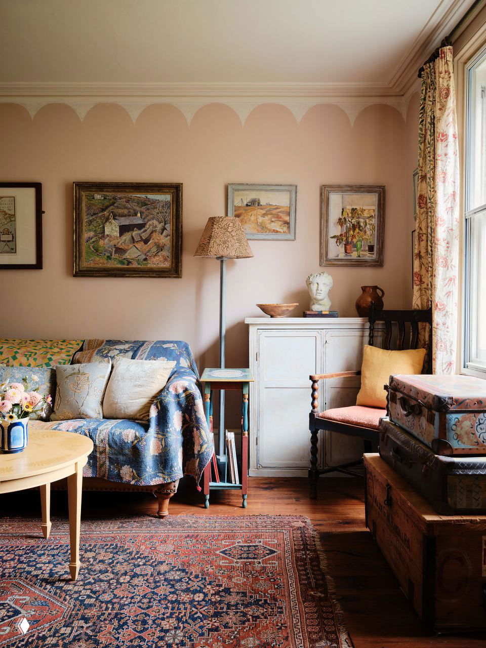 Светлая комната с диваном, узорным пледом, настенным искусством и сундуками у окна — пастельные тона и винтажные акценты.