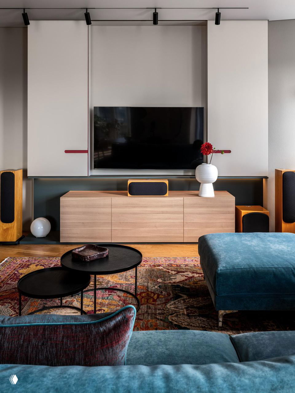 ТВ-зона: белые панельные шкафы, встроенный телевизор, вазa с цветами на тумбе и насыщенный декоративный ковёр на полу.