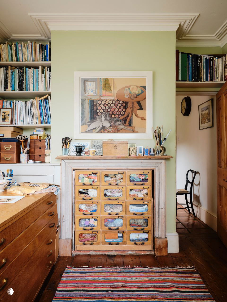 Рабочая зона и книжные полки: деревянные шкафы, маленькие выдвижные ящики с тканями и картина над камином, аккуратный художественный беспорядок.