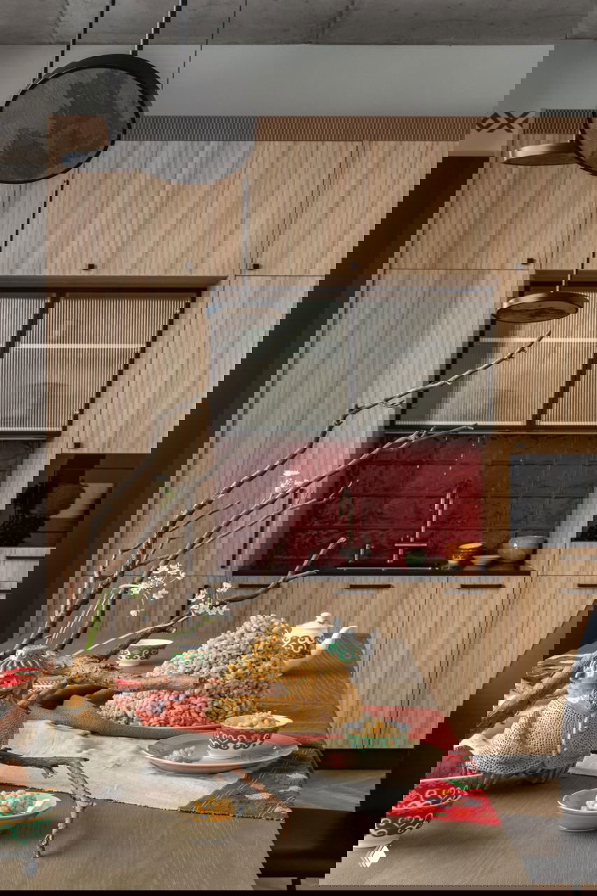 Ближний план стола с композициями: ветки, декоративные скульптуры и блюда на фоне деревянной мебели и красной плитки в офисной кухне.