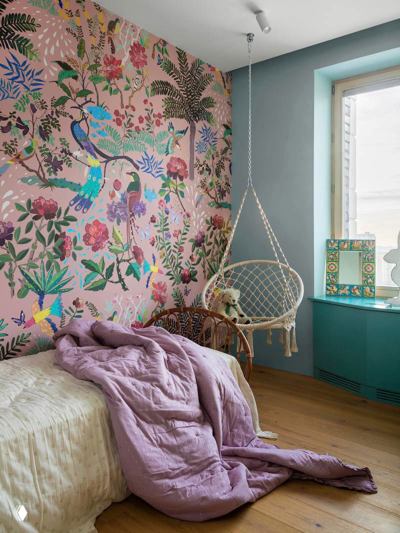 Детская или спальня с розовой стеной, расписанной птицами и растительными мотивами, подвесное плетёное кресло и мягкое постельное бельё на переднем плане.