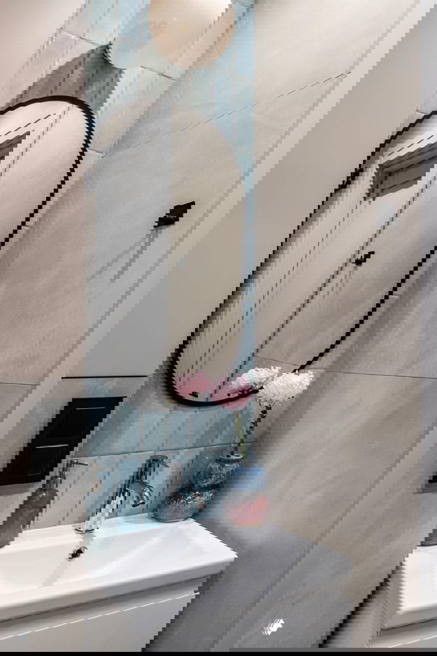 Умывальник в ванной с овальным зеркалом, голубой декоративной плиткой за зеркалом и керамической вазой с цветами — небольшие декоративные решения в санузле.