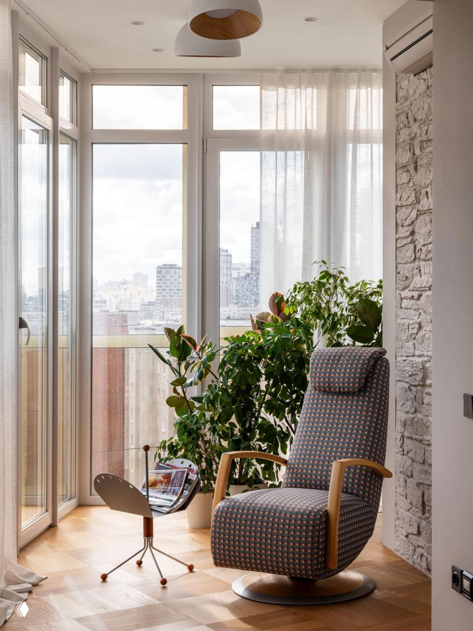 Уютный уголок у панорамного окна: кресло-реклайнер в узорной ткани, журнальный держатель и крупные комнатные растения на фоне городского пейзажа.