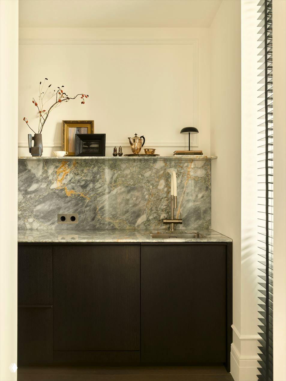 Небольшая мини-кухня с мраморной стеновой панелью и темными фасадами, аккуратные предметы декора и минималистичный смеситель в интерьере квартиры.