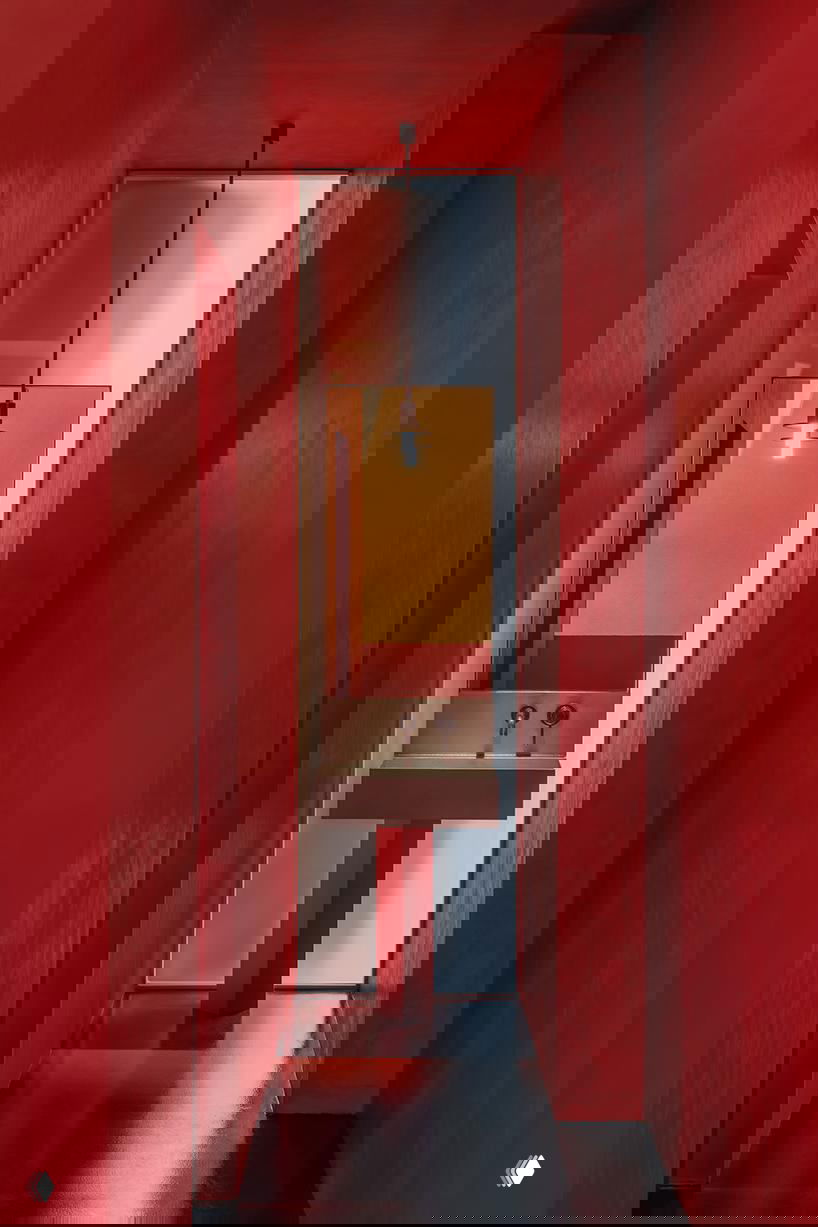 Узкий коридор с насыщенно-красными стенами, простая раковина и подвесной светильник на фоне тонкой вертикальной щели-окна, минималистичный декор.