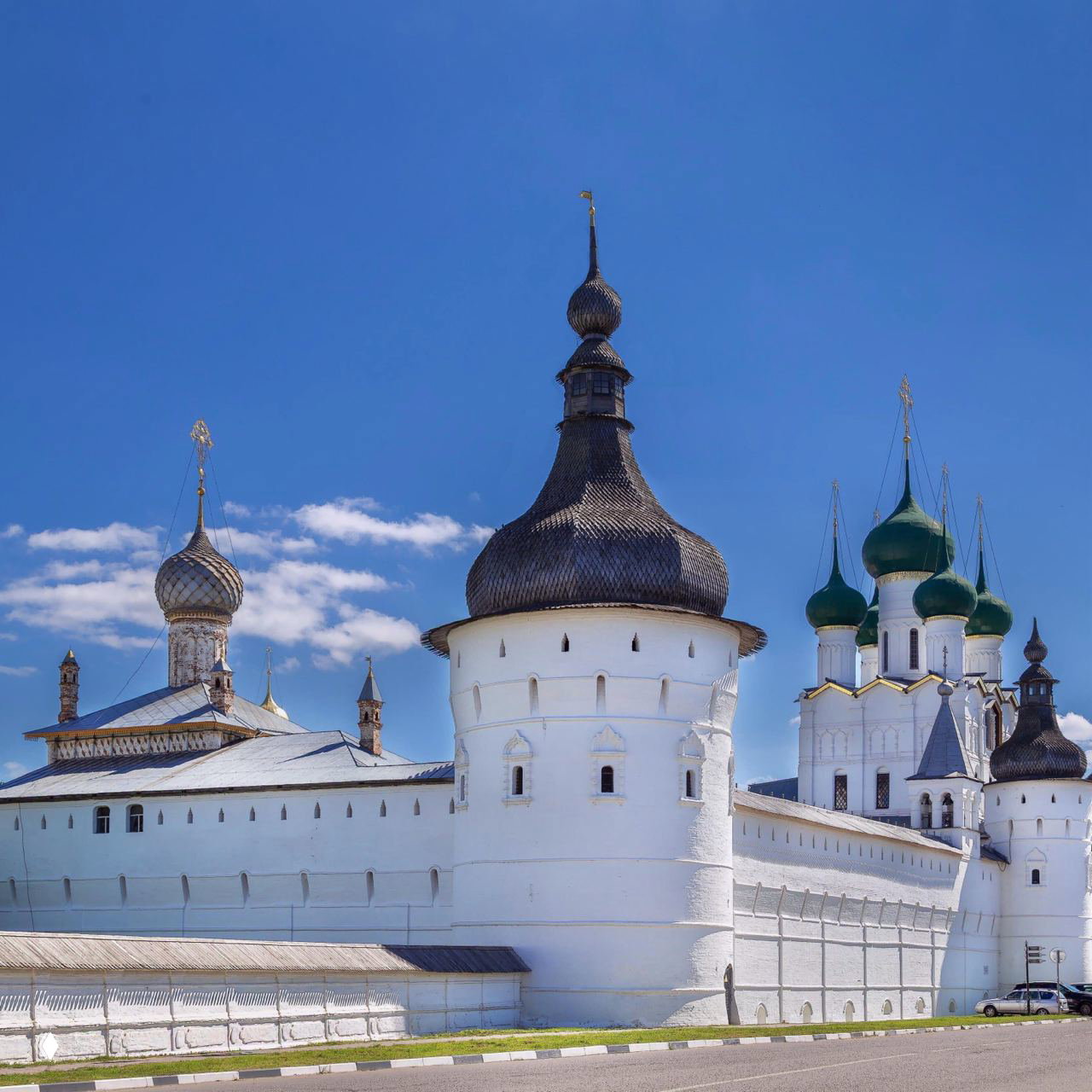 Массивная белая башня и фрагменты стен крепости с зелёными куполами церкви на фоне ясного синего неба и городской дороги у подножия.