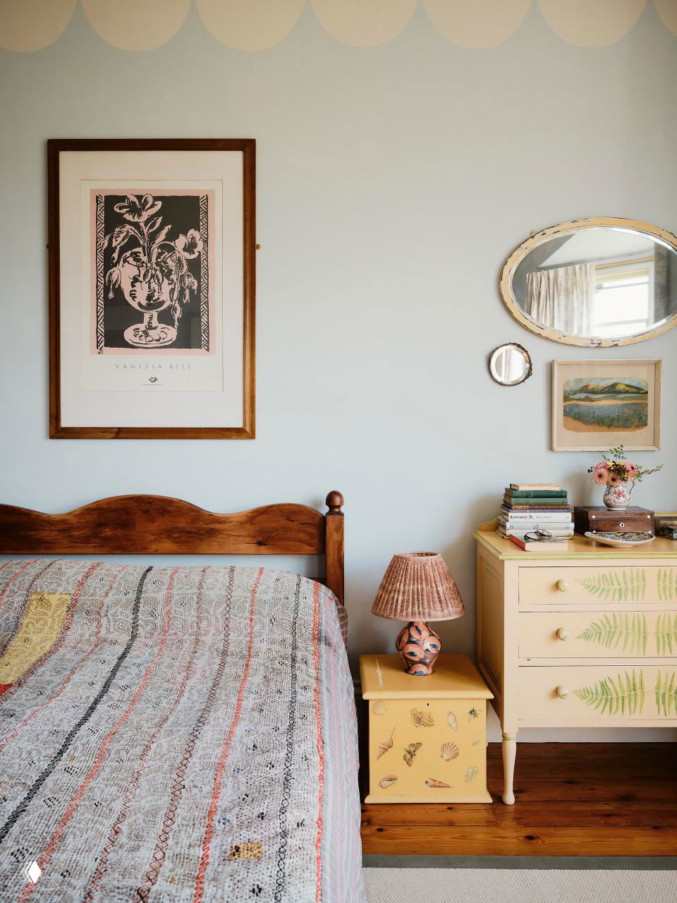 Спальня с деревянной кроватью, тумбочкой с расписным декором и лампой в тёплых тонах; постельные текстуры и пастельные стены.