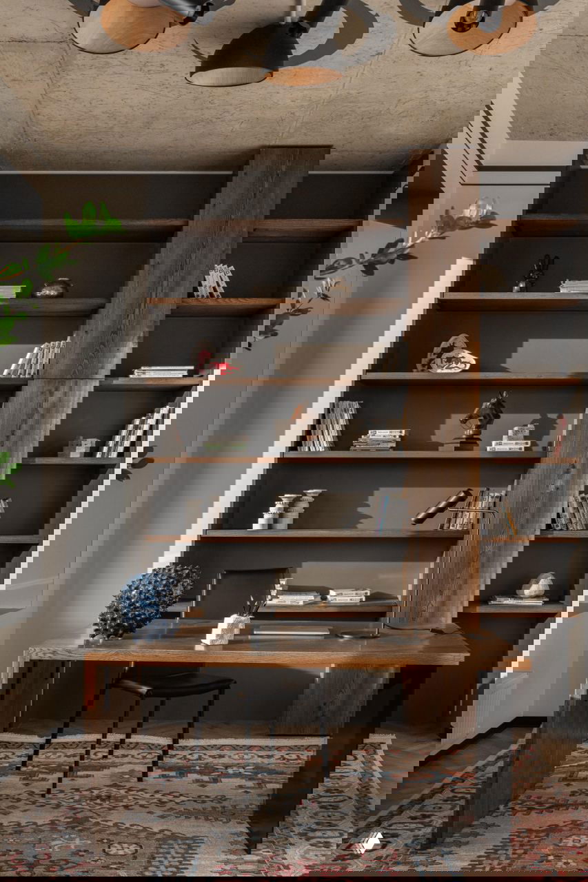 Рабочее место у стенки с высокими деревянными полками: стол, лампа, декоративные книги и предметы на фоне деревянного книжного стеллажа.