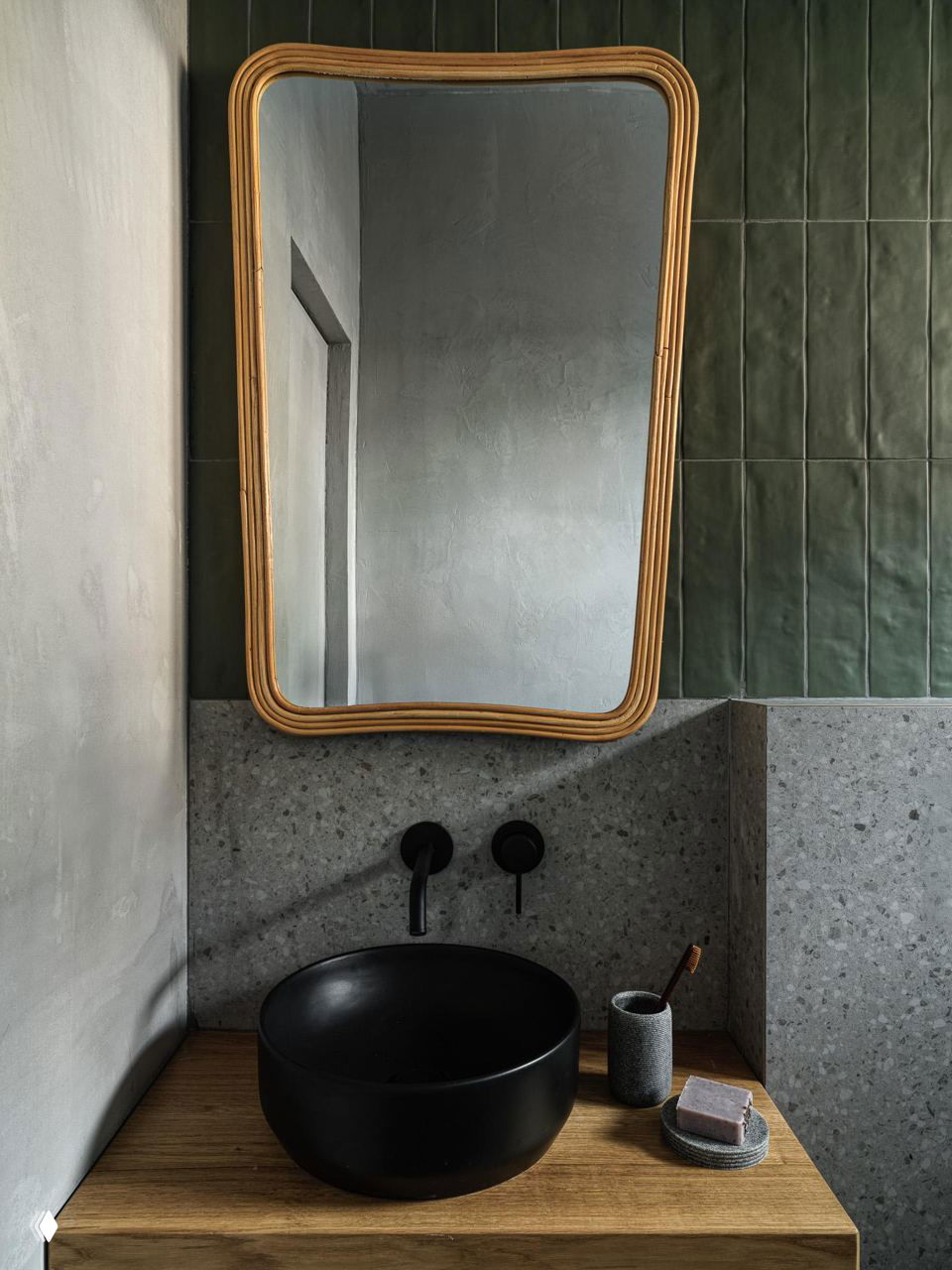 Раковина-чаша чёрного цвета на деревянной тумбе, овальное золочёное зеркало и фактурная зелёная плитка с бетонной отделкой стен в ванной комнате.