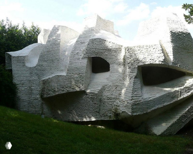 Крупный план белой фактурной кладки и резких геометрических выемок фасада дома-скульптуры, окружённого травой и деревьями.