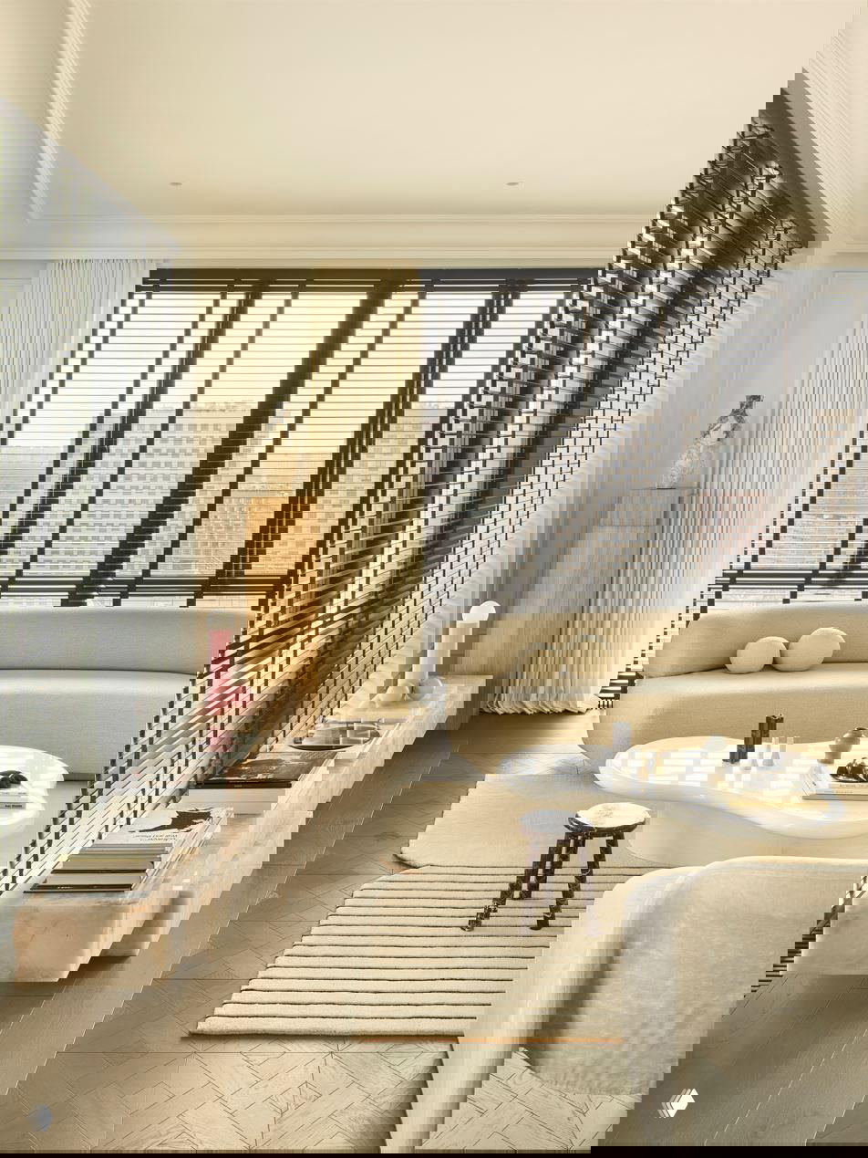 Светлая гостиная с низким изогнутым диваном бежевого цвета, широкие окна в черных рамах и прозрачные жалюзи, мягкий дневной свет заливает пространство.