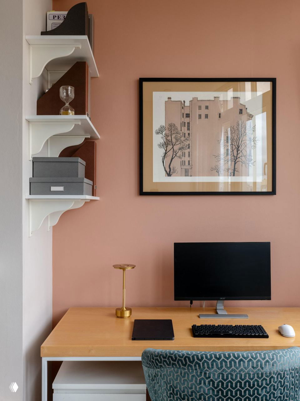 Рабочее место в квартире: стол с монитором и клавиатурой, настольные полки и декоративная картина на фоне персиковой стены, организованная зона для работы.