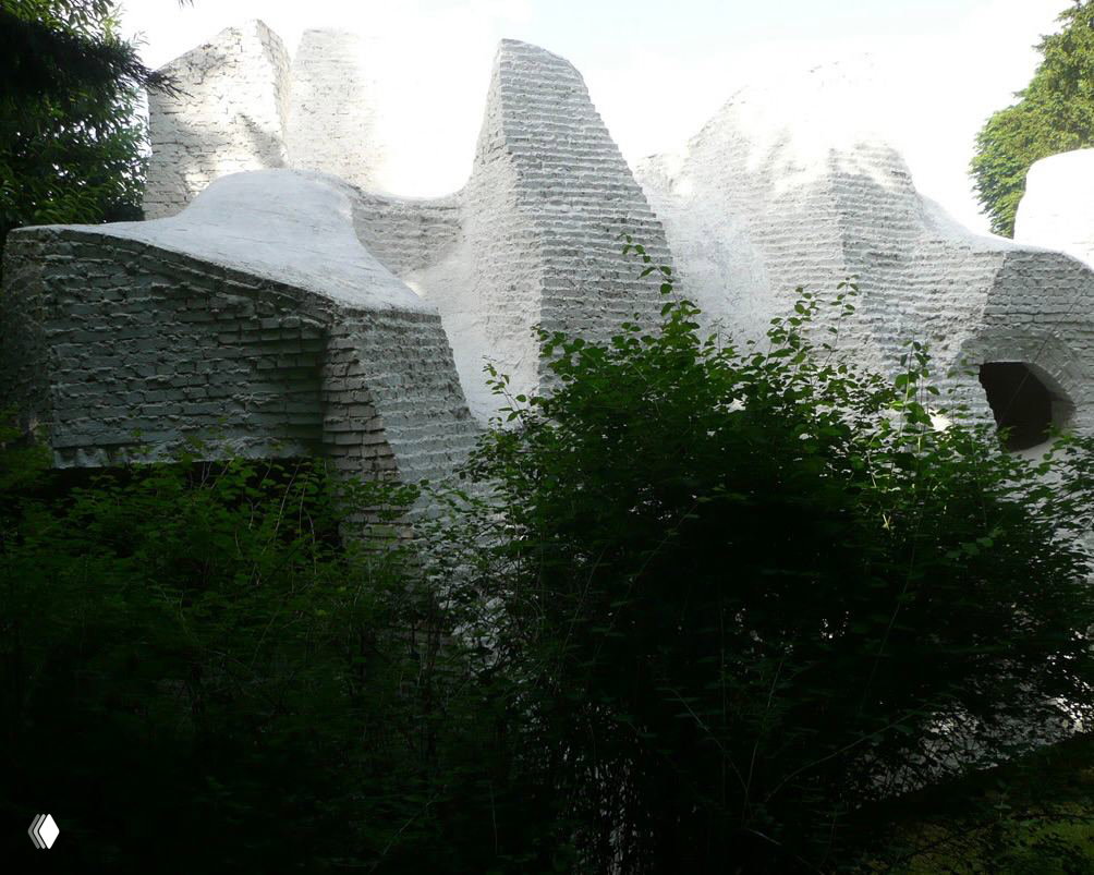 Вид сбоку на сложную композицию белых объёмов дома, частично скрытую зеленью, с характерными округлыми проёмами и нишами.