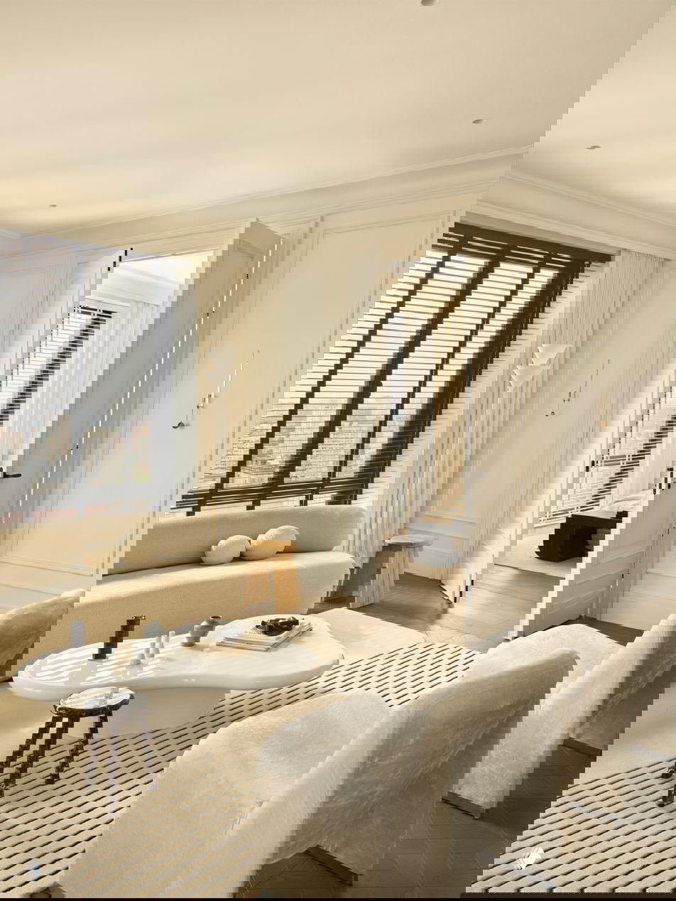 Интерьер гостиной с открытым проходом в спальню, классические молдинги стен, нейтральная палитра и деревянный паркет темного тона создают элегантную атмосферу.