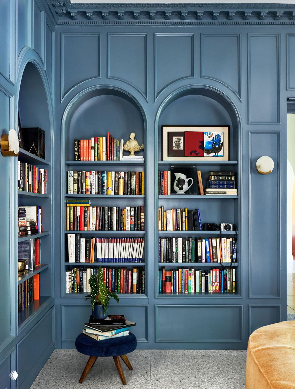 Кабинет с встроенными книжными полками в нишах, тёмно‑синие стеновые панели, множество книг и бархатный небольшой табурет у полок.