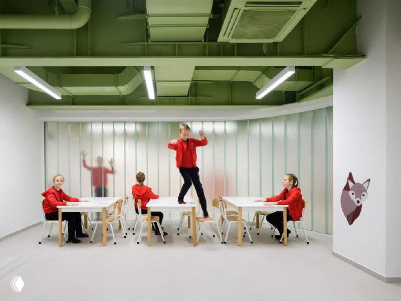 Учебная зона рядом с наклонной колонной: столы, стулья и естественное освещение в просторном светлом интерьере школьного помещения.