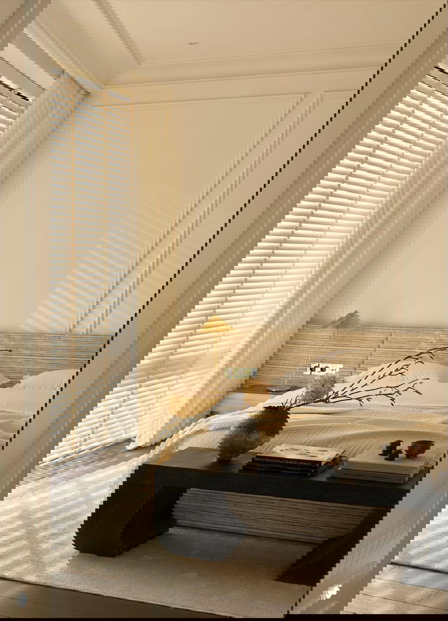 Спальня в теплых светлых тонах: низкое изголовье, текстильные жалюзи у окна, декоративная ваза на столике и мягкое естественное освещение.