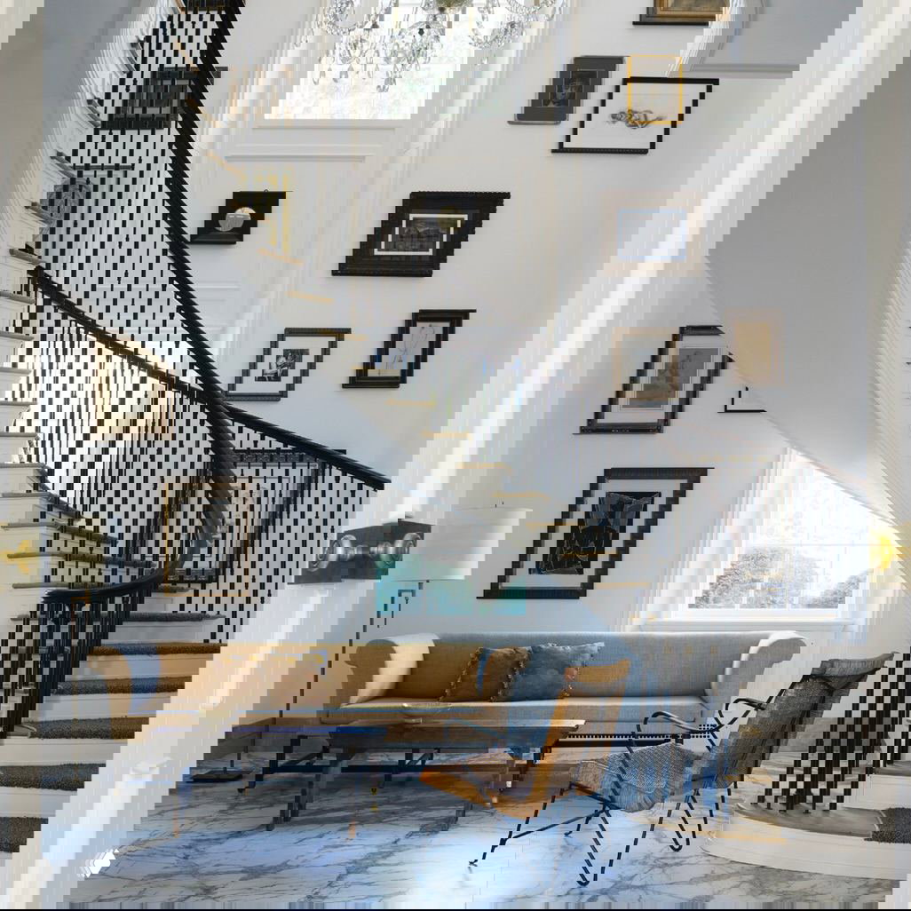 Витая лестница в холле особняка, высокие окна, мраморный пол у входа и элегантный диван с креслом на фоне галереи рам на стене