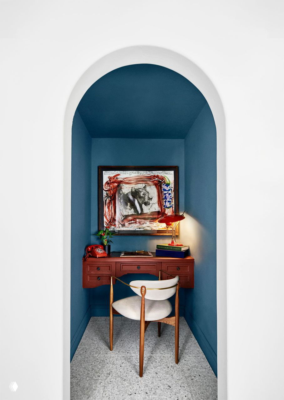 Арочная ниша, где расположен красный письменный стол, настольная лампа и стул — компактное рабочее место в глубоком синем оттенке.