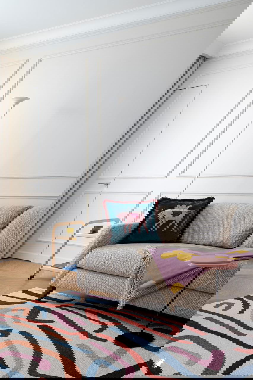 Уютный фрагмент гостиной: текстурный светлый диван, декоративная подушка с рисунком, разноцветный плед и крупный абстрактный ковёр на паркетном полу.