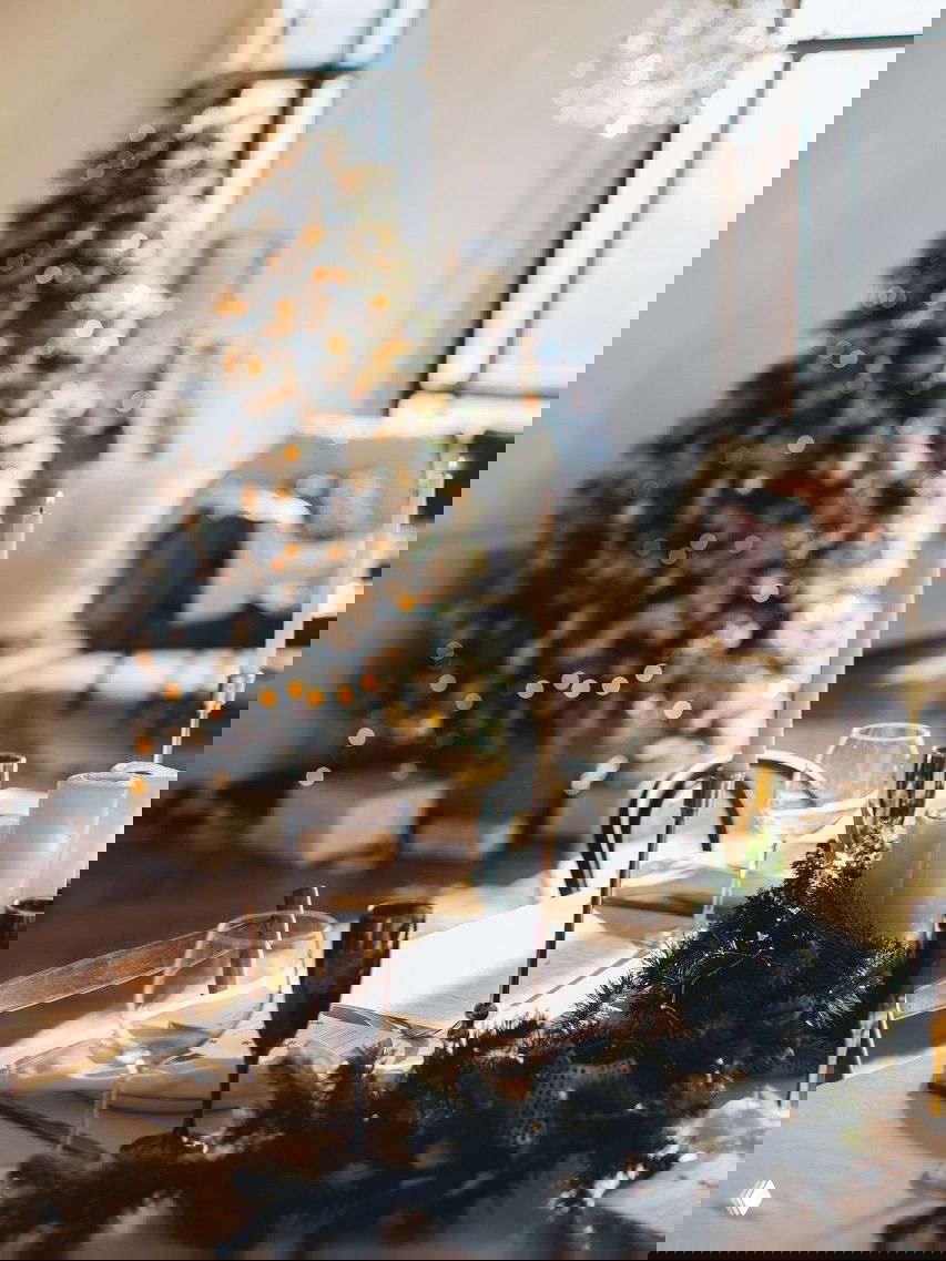 Сервированный праздничный стол со свечами, бокалами и гирляндой на фоне размытой ёлки и мягкого дневного света — атмосферная новогодняя сервировка.