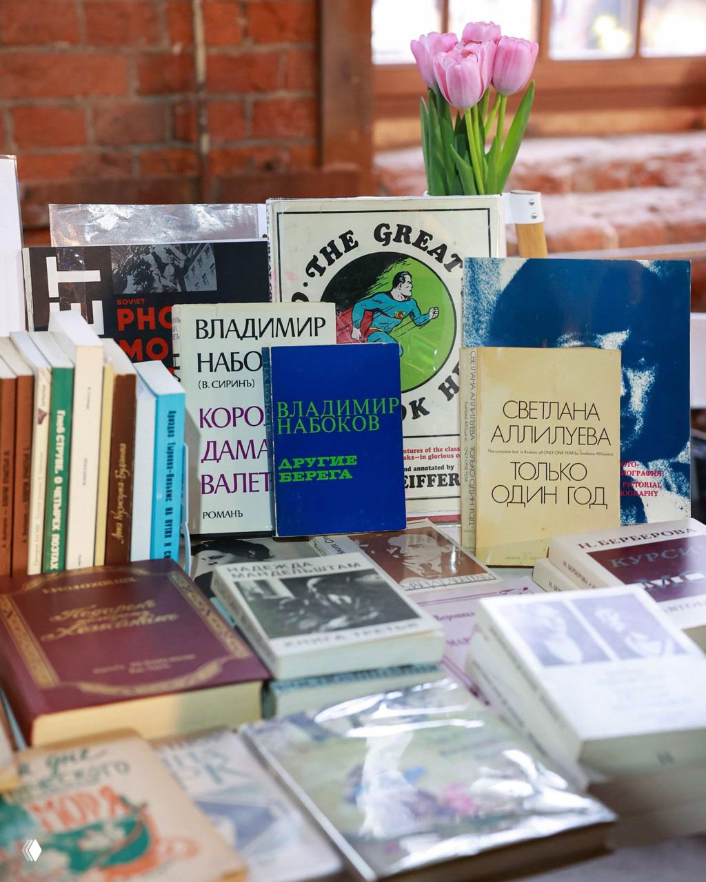 Стол с книгами на ярмарке: подборка изданий, в том числе Владимир Набоков, на фоне тюльпанов и кирпичной стены.