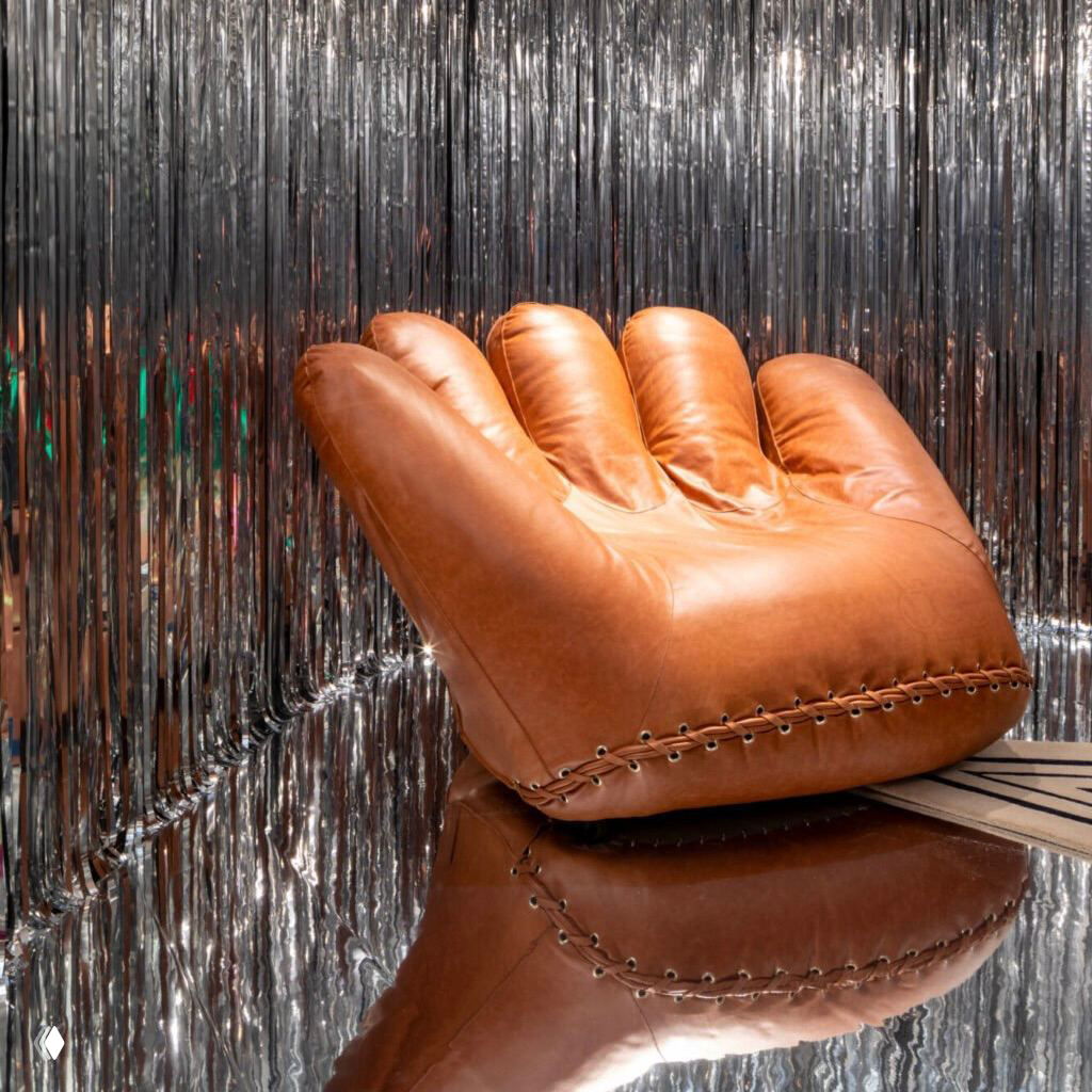 Кресло Joe установлено на зеркальной поверхности на фоне фольгированной завесы; видно отражение изделия и акцент на силуэте перчатки.