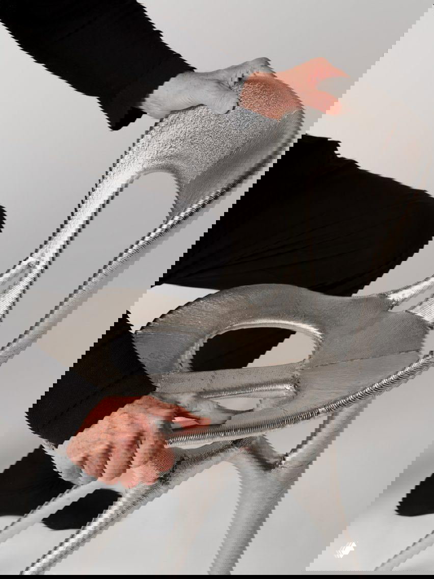 Детальный план рук, рассматривающих металлическую 3D-печатную конструкцию сиденья; заметна текстура и плавные переходы в форме изделия.