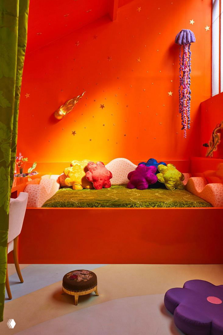 Спальная ниша с оранжевой стеной и множеством ярких декоративных подушек‑цветов на кровати — сказочный, театральный декор в интерьере Uchronia.