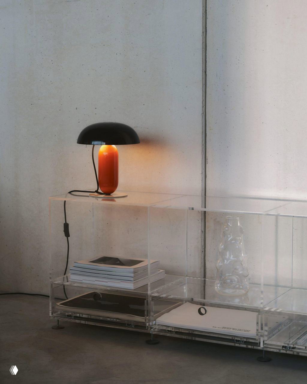 Чёрно-оранжевый вариант лампы Gambosa на прозрачной мебели у бетонной стены; дизайн без видимых соединений и эффект визуального баланса деталей.