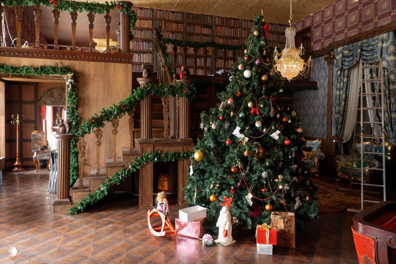 Интерьер усадьбы с украшенной рождественской ёлкой, библиотечными полками и лестницей, праздничные гирлянды и подарки на полу.