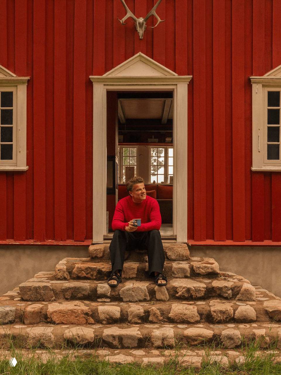 Бьярке Ингельс сидит на каменных ступенях у входа в красный коттедж, держит кружку, на заднем плане видно внутреннее помещение и окно с видом на сад.