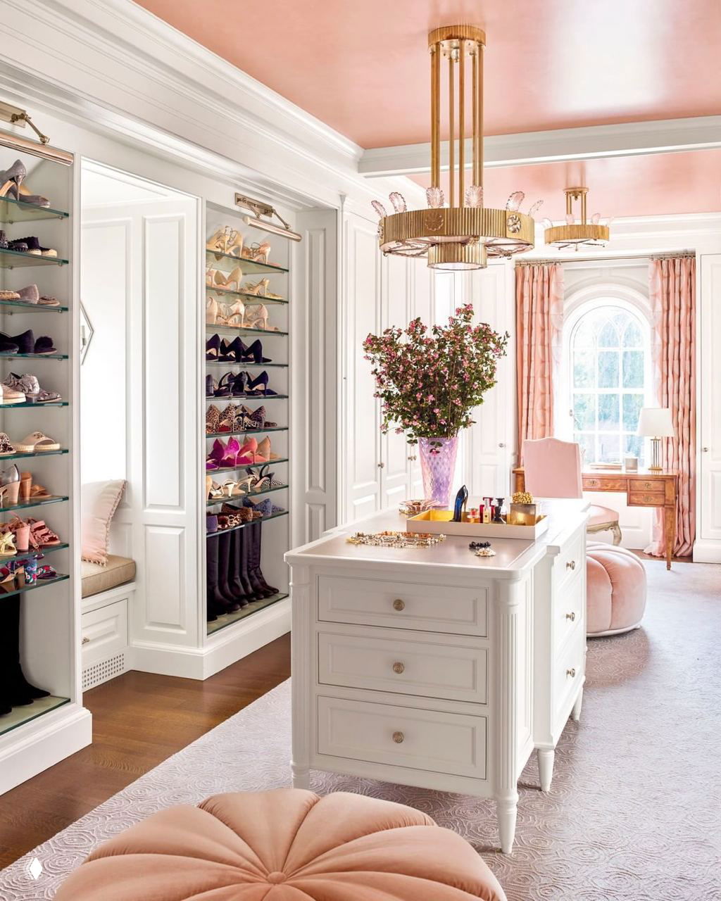 Роскошная гардеробная в пастельных тонах: остров с ящиками, стенды для обуви, мягкие пуфы и золотистая люстра создают интерьерную комнату-бутик.