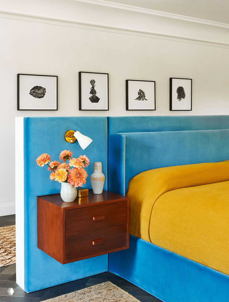 Спальня с ярким велюровым голубым изголовьем, желтым покрывалом, плавающей прикроватной тумбой и вазой с цветами для акцента цвета.