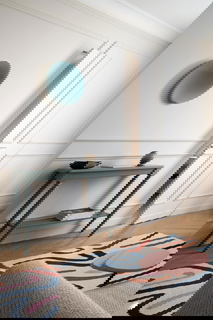 Фрагмент прихожей с зелёной консолью, декоративной тарелкой на стене и краем яркого ковра на паркетном полу — минималистичный и продуманный интерьер.