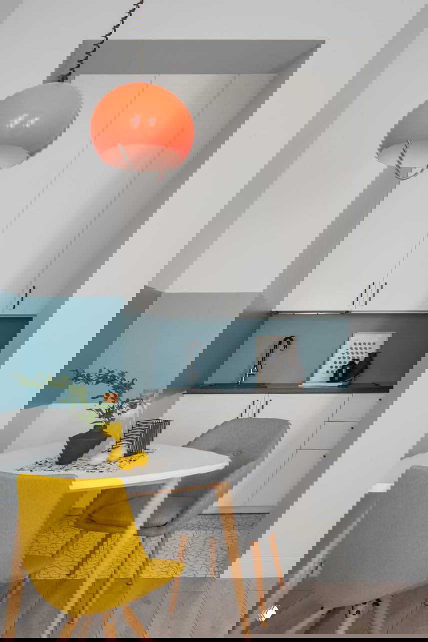 Небольшая нишевая кухня с голубым фартуком, яркой оранжевой подвесной лампой и желтым стулом у обеденного столика в скандинавском стиле.