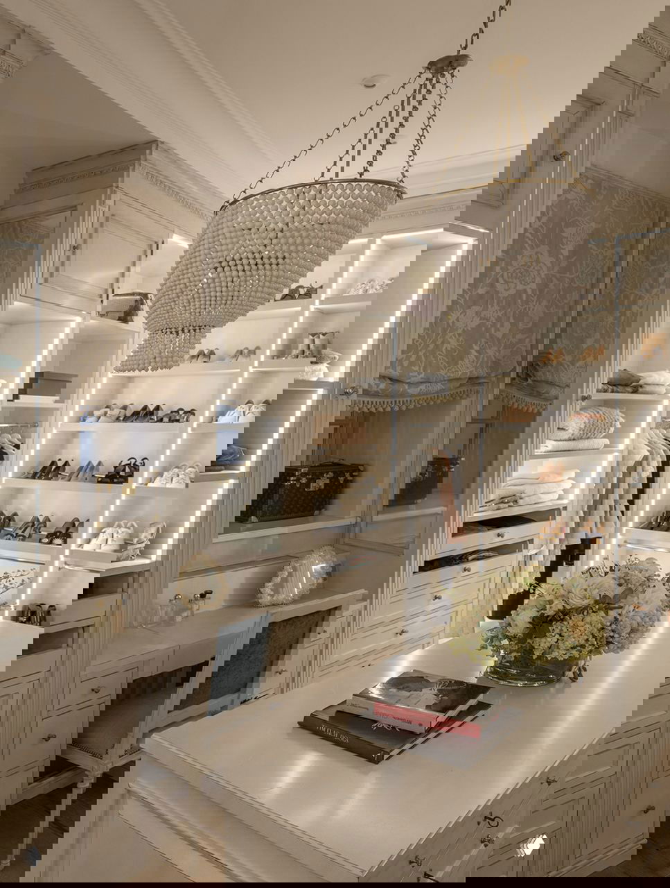 Классическая светлая гардеробная с декоративной лепниной, встроенным освещением полок, туалетным столиком и центральной столешницей с цветами.