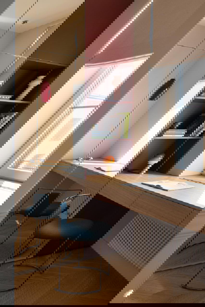 Рабочая зона у окна: длинная деревянная столешница, открытые полки бордового цвета, зеркальные фасады и две тёмно-бирюзовые табуретки для компактного места работы.