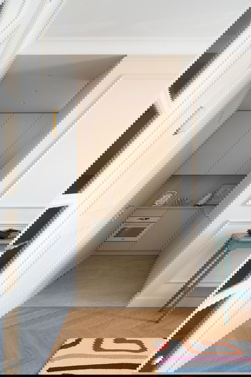 Небольшая компактная кухня в светло-бежевых тонах с встраиваемой техникой и аккуратной рабочей зоной, плавно переходящая в гостиную с паркетом.