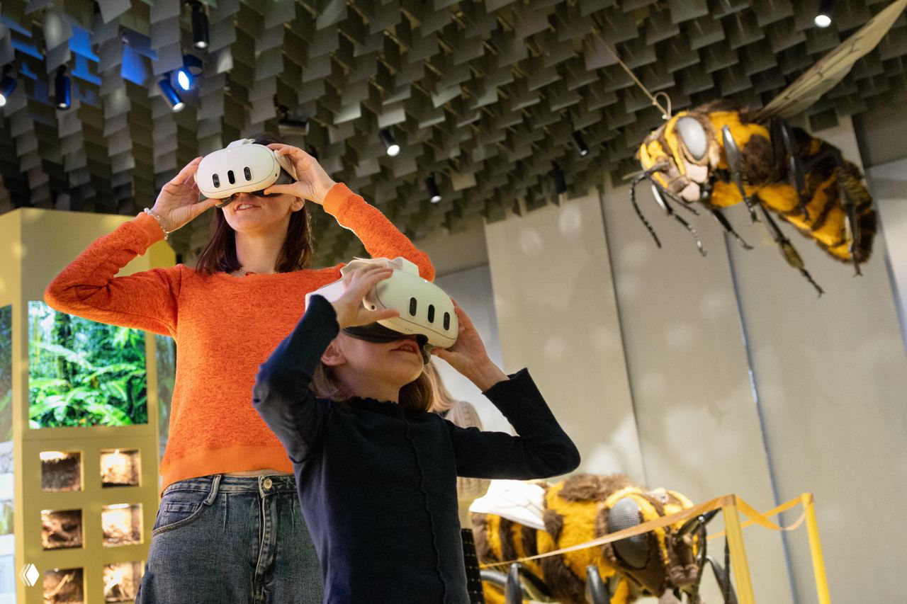 Посетители в очках виртуальной реальности на интерактивной выставке в биокластере; большой макет насекомого на заднем плане.